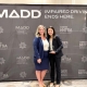 Two women in business attire smile while posing together in front of a MADD (Mothers Against Drunk Driving) event backdrop that reads, "Impaired Driving Ends Here." The woman on the right holds a glass award, recognizing her achievement, as they stand side by side at the awards event.