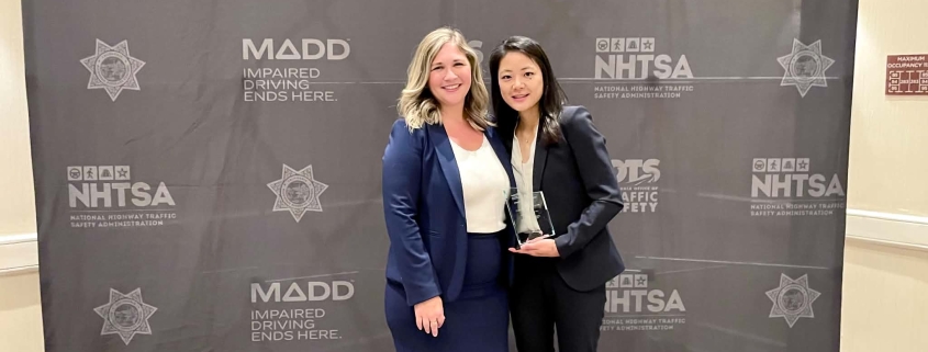 Two women in business attire smile while posing together in front of a MADD (Mothers Against Drunk Driving) event backdrop that reads, "Impaired Driving Ends Here." The woman on the right holds a glass award, recognizing her achievement, as they stand side by side at the awards event.