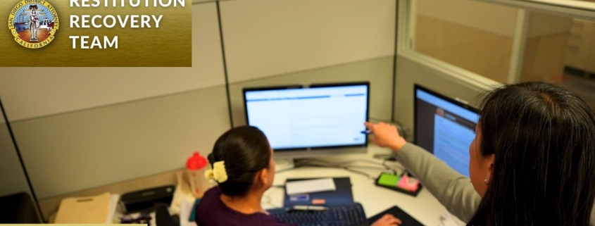 Two members of the San Diego District Attorney's Restitution Recovery Team working together at a desk, reviewing information on computer monitors in an office cubicle.