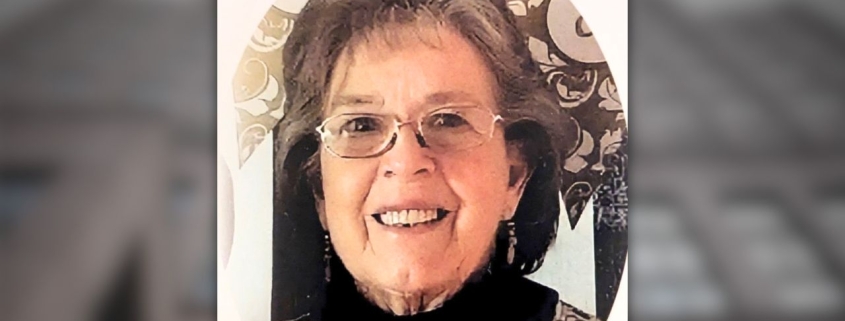 Smiling woman with short dark hair and glasses, wearing dangling earrings and a patterned top, photographed against a light background with a decorative circular frame.