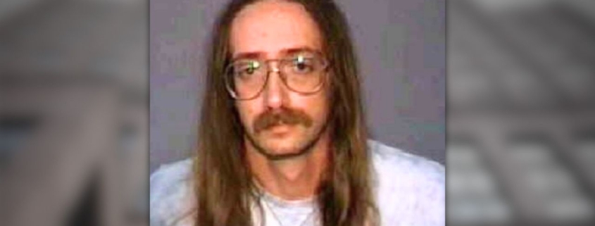 Retrato de medio cuerpo de un hombre con cabello largo y castaño y bigote, usando gafas redondas y una camiseta de color claro, frente a un fondo gris liso; el texto indica: “Foto de la víctima: Cecil Zachary Jr.”