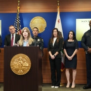 A group of officials and uniformed officers stand in front of a podium with a seal, American flags, and a blue backdrop during a formal press event.