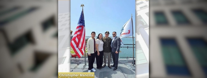 A group of four people posing outdoors near a chain-link fence with an American flag on the left and a U.S. Army flag on the right under a clear blue sky.