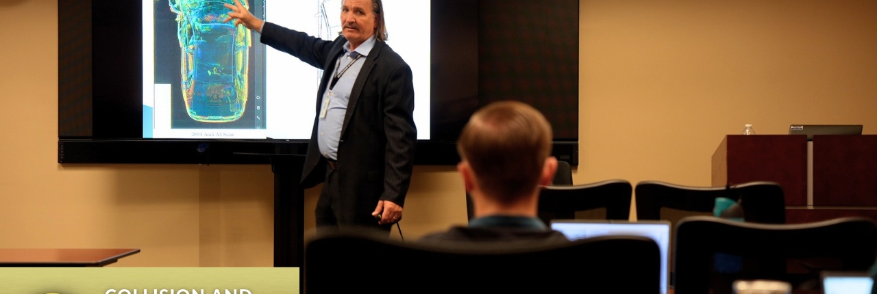 A man stands in front of a screen projector showing the image of a car to an audience in a classroom training room.