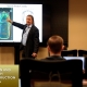 A man stands in front of a screen projector showing the image of a car to an audience in a classroom training room.