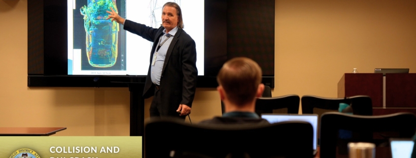 A man stands in front of a screen projector showing the image of a car to an audience in a classroom training room.