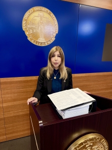 DA Summer Stephan stands behind a podium in front of the DA Seal with a large binder with a lot of documents.