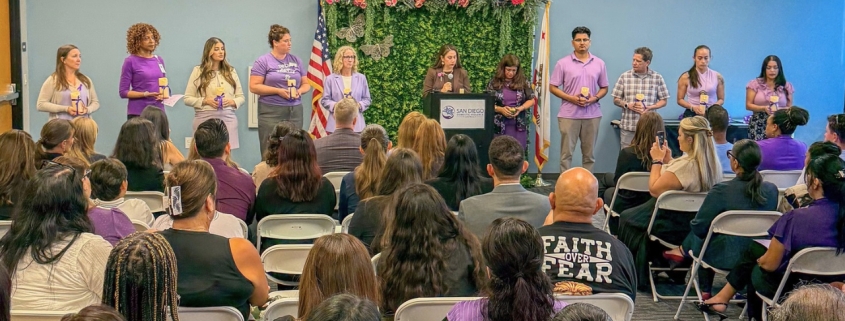 A group of people in purple shirts stand with candles in front of a floral backdrop, while an audience sits in rows facing the podium and speaker.