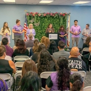 Un grupo de personas con camisas moradas sostiene velas frente a un fondo floral, mientras el público se sienta en filas frente al podio y al orador.