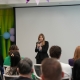 Una mujer vestida con un saco oscuro habla por un micrófono durante un pequeño evento en un espacio interior, mientras los asistentes sentados la observan. Detrás de ella hay una pantalla de proyección en blanco, un fondo decorativo estilo bambú verde y globos azules y morados.