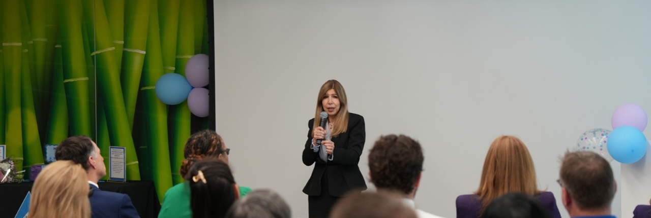 A woman in a dark blazer speaks into a microphone at a small indoor event while seated attendees watch; behind her are a blank projection screen, green bamboo-style backdrop, and blue and purple balloons.
