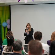 A woman in a dark blazer speaks into a microphone at a small indoor event while seated attendees watch; behind her are a blank projection screen, green bamboo-style backdrop, and blue and purple balloons.