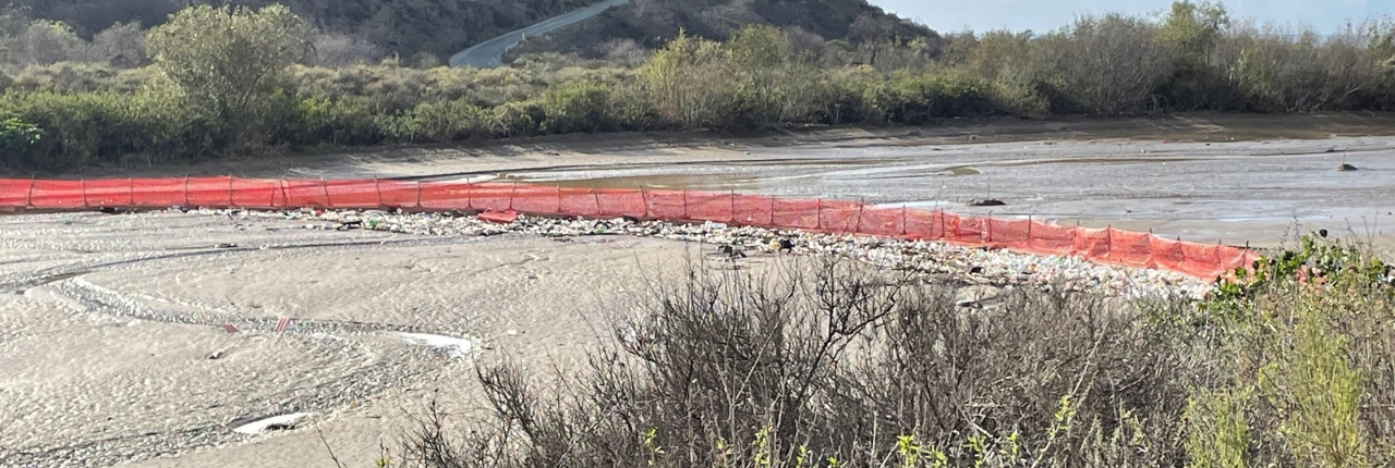A wide riverbank with gravel and shrubs in the foreground, a red barrier wall lining the water, and forested hills and mountains under a partly cloudy sky.
