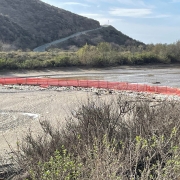 A wide riverbank with gravel and shrubs in the foreground, a red barrier wall lining the water, and forested hills and mountains under a partly cloudy sky.