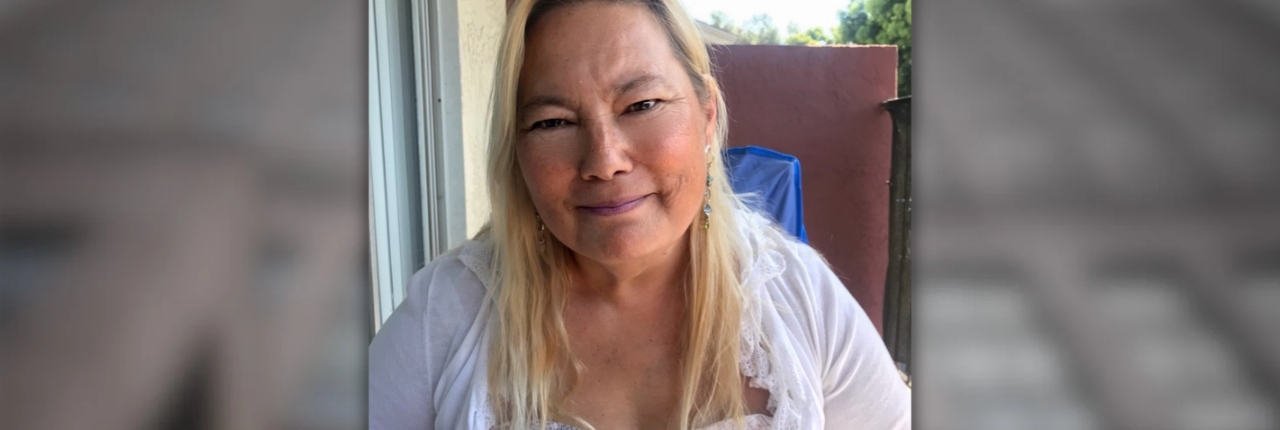 Portrait of a woman with shoulder-length blonde hair wearing a white lace-trim top, smiling gently in an outdoor patio setting with a blue tarp and red wall in the background.