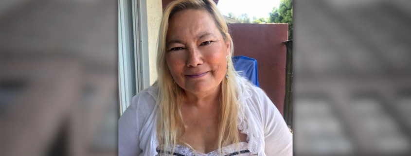Portrait of a woman with shoulder-length blonde hair wearing a white lace-trim top, smiling gently in an outdoor patio setting with a blue tarp and red wall in the background.