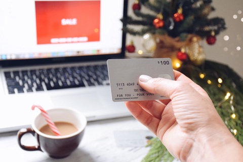 Mano de mujer con tarjeta de credito a punto de hacer compras por Internet a traves de su computadora portatil, junto con una taza de cafe y el arbolito de Navidad al fondo.